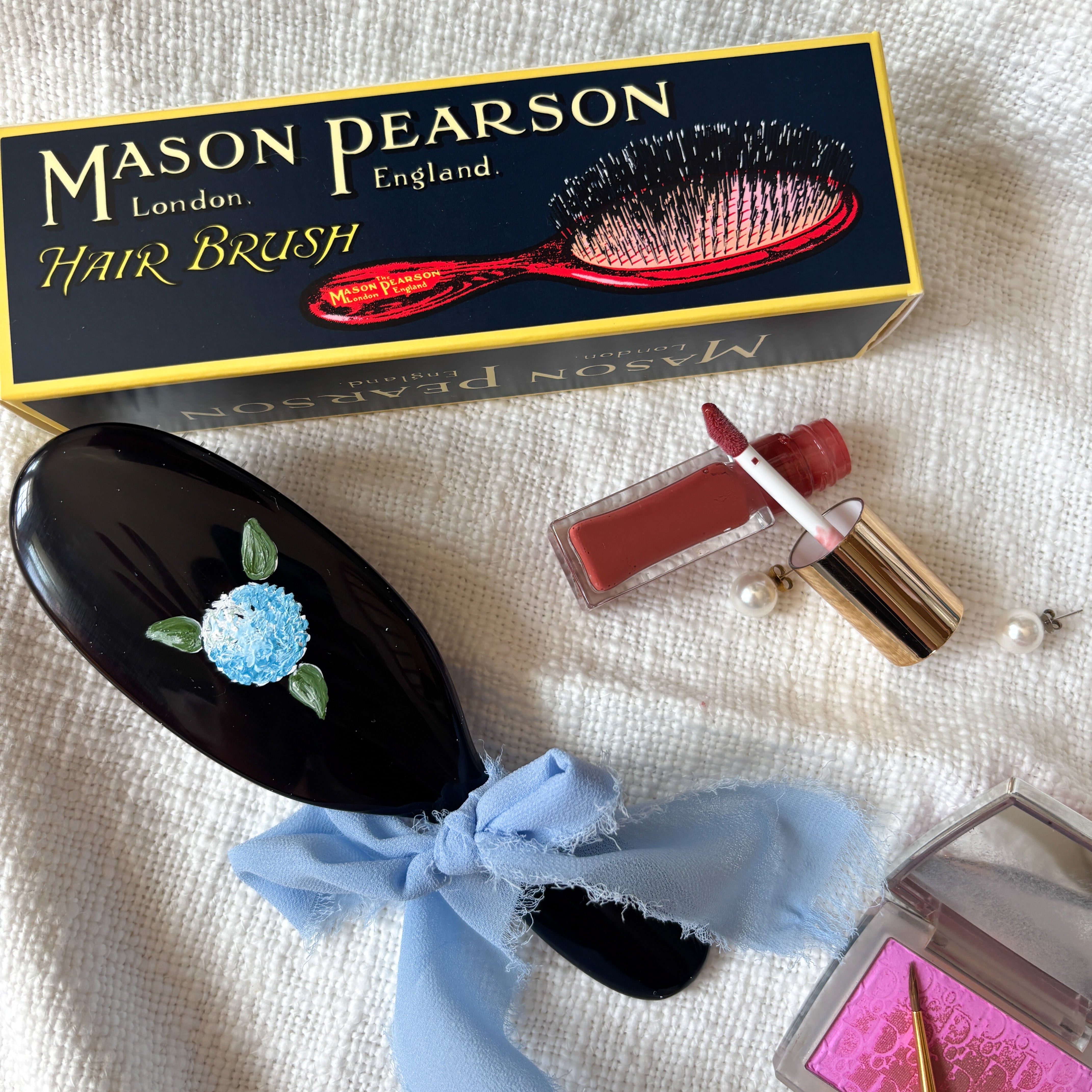 Mason Pearson hairbrush with decorative bow, lipstick, and makeup palette on a textured surface