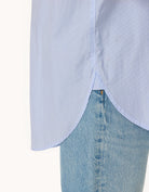 Close-up of the high-low hem detail of a light blue shirt and jeans on a white background
