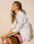 Woman wearing a white button down shirt and multicolor striped skirt sitting against a beige wall.