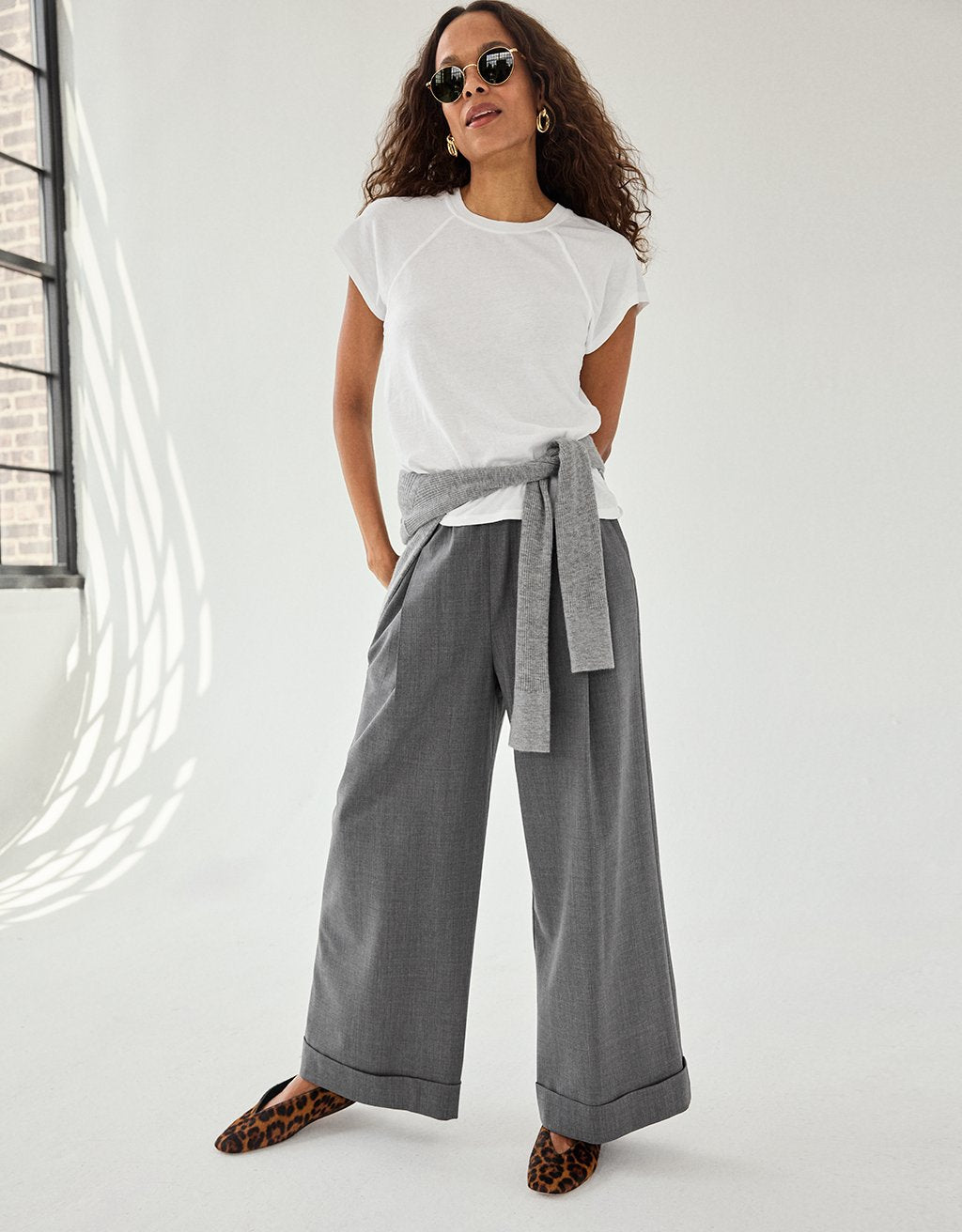 Woman wearing a white raglan t-shirt and gray wide-leg pants with a gray sweater tied around her waist against a white background with an exposed window