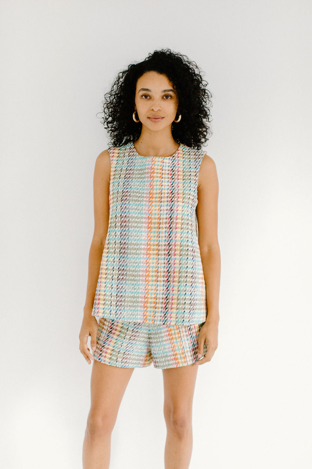 A woman with olive skin and short, curly black hair staring directly at the camera. She's wearing gold hoop earrings and a matching wide cut tank top and shorts in a colorful plaid pattern.