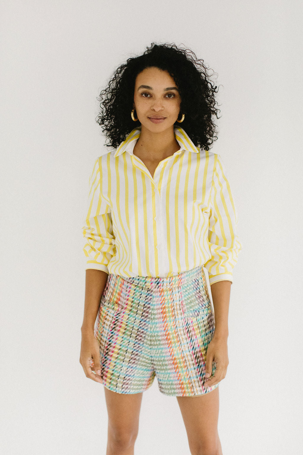 A woman with olive skin and short, black, curly hair staring directly at the camera with a small smile. She's wearing chunky gold hoop earrings, a white and yellow striped button down shirt pushed up to the elbows and a pair of colorful plaid shorts.