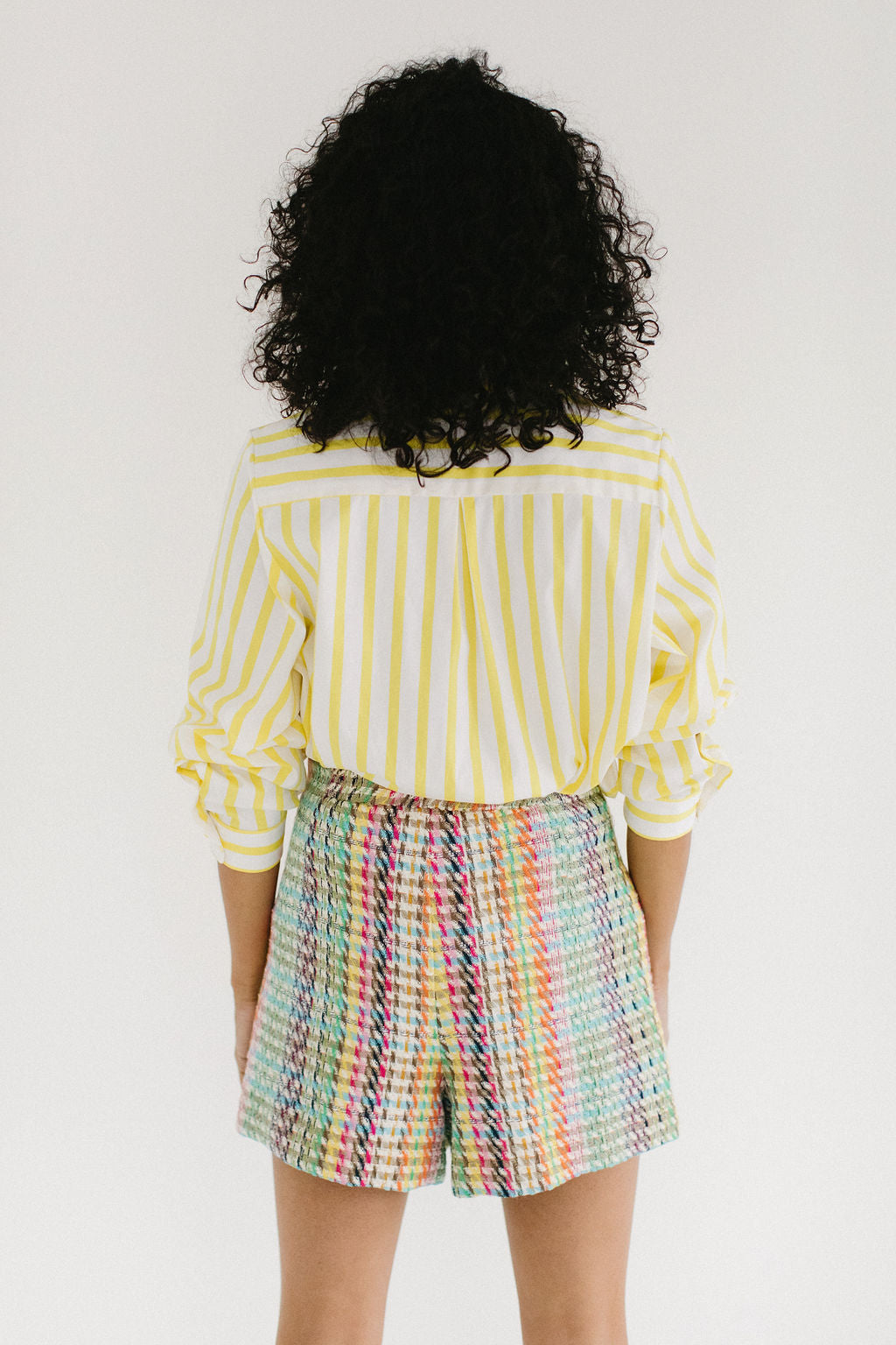 A back view of a woman with olive skin and short, black, curly hair staring directly at the camera with a small smile. She's wearing chunky gold hoop earrings, a white and yellow striped button down shirt pushed up to the elbows and a pair of colorful plaid shorts.