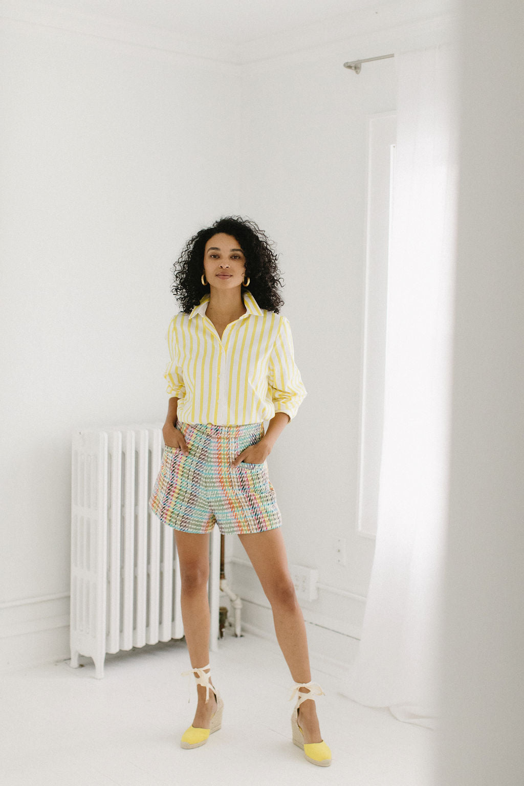 A full body shot of a woman with olive skin and short, black, curly hair staring directly at the camera with a small smile. She's wearing chunky gold hoop earrings, a white and yellow striped button down shirt pushed up to the elbows and a pair of colorful plaid shorts. She's wearing a pair of yellow espadrille heeels that are tied around her ankles. You can see a window and a white radiator in the background of the image.