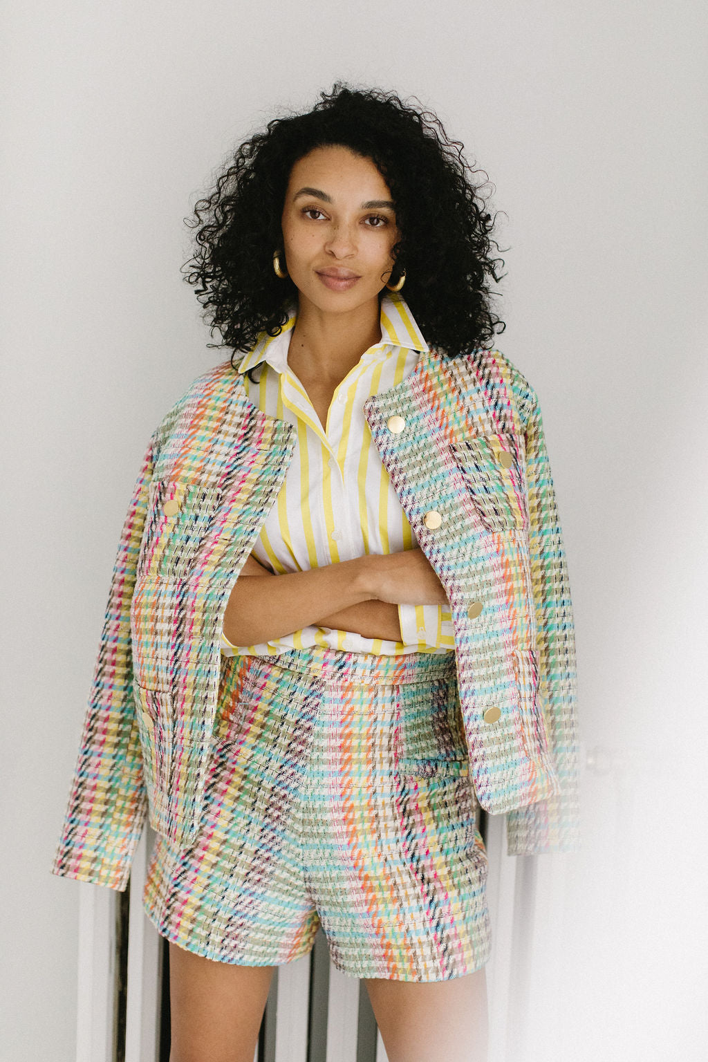 A woman with olive skin and short, black, curly hair staring directly at the camera with a small smile. She's wearing chunky gold hoop earrings, a white and yellow striped button down shirt pushed up to the elbows and a pair of colorful plaid shorts. She had her arms crossed around her waist and a matching colorful plaid jacket draped around her shoulders.