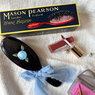 Mason Pearson hairbrush with decorative bow, lipstick, and makeup palette on a textured surface
