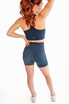 A view of the back of a woman with red, curly hair wearing a navy sports bra and biker shorts set and white sneakers. She has her left hand on her hip and her right hand adjusting her ponytail.