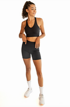 A full body shot of a woman gazing away from the camera and adjusting the waistband of her black biker shorts. She's wearing a vneck black sports bra and high cut nike socks and Nike sneakers. 