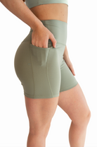 Side close up view of a woman wearing a sage green workout set with her hand tucked into the side pocket of the biker shorts.