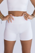 Close up of a woman wearing lots of stacked bracelets and rings with her hands on her hips. She's wearing a white workout set with the focus on the biker shorts.