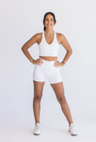 Straight on view of a woman wearing a white workout set (Vneck sports bra and biker shorts). She's smiling at the camera and posing with her left leg straightened out to her left side.