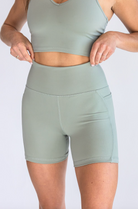 Closeup of the midsection of a woman wearing a sage green sports bra and biker shorts set. She's adjusting the waistband of the shorts with both hands.