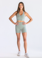Head on view of a woman wearing a sage green workout set (Vneck sports bra and biker shorts set). She has long brown hair that's straight and hanging over her shoulders.