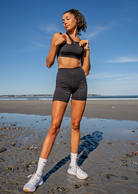 A woman on the beach in a black workout set of a sports bra and biker shorts with high cut socks and sneakers. She's looking down toward the ground turning her head with her eyes closed basking in the sun.